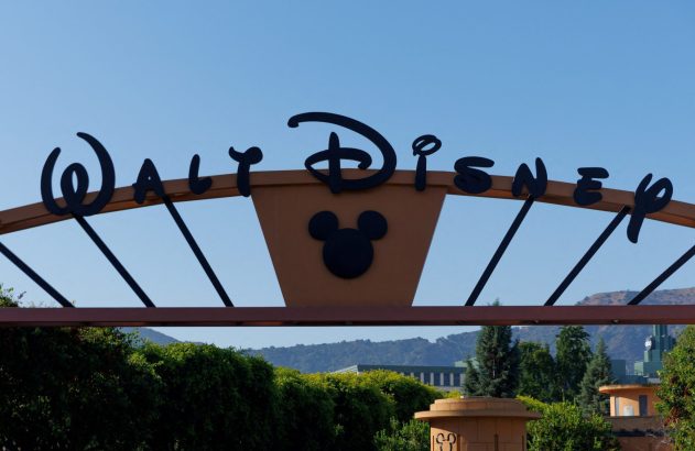 Sinal de entrada da Walt Disney com logotipo e silhueta do Mickey Mouse, sob céu azul.
