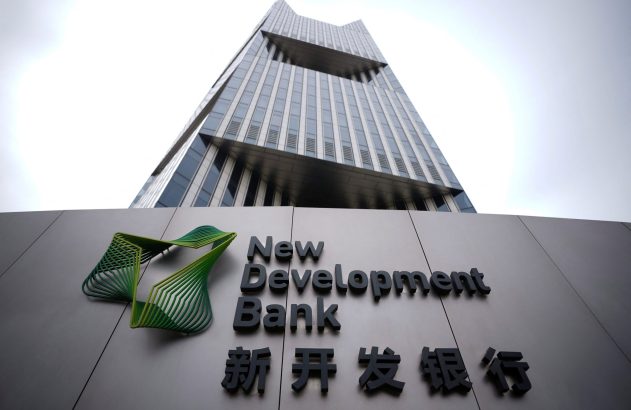 Prédio moderno. Letreiro cinza com logo verde e 'New Development Bank' em inglês e chinês na frente.