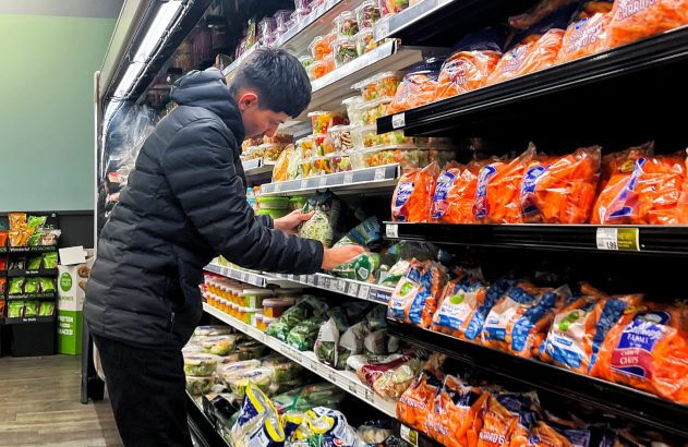 Homem de jaqueta escura escolhendo vegetais embalados em prateleira de supermercado.