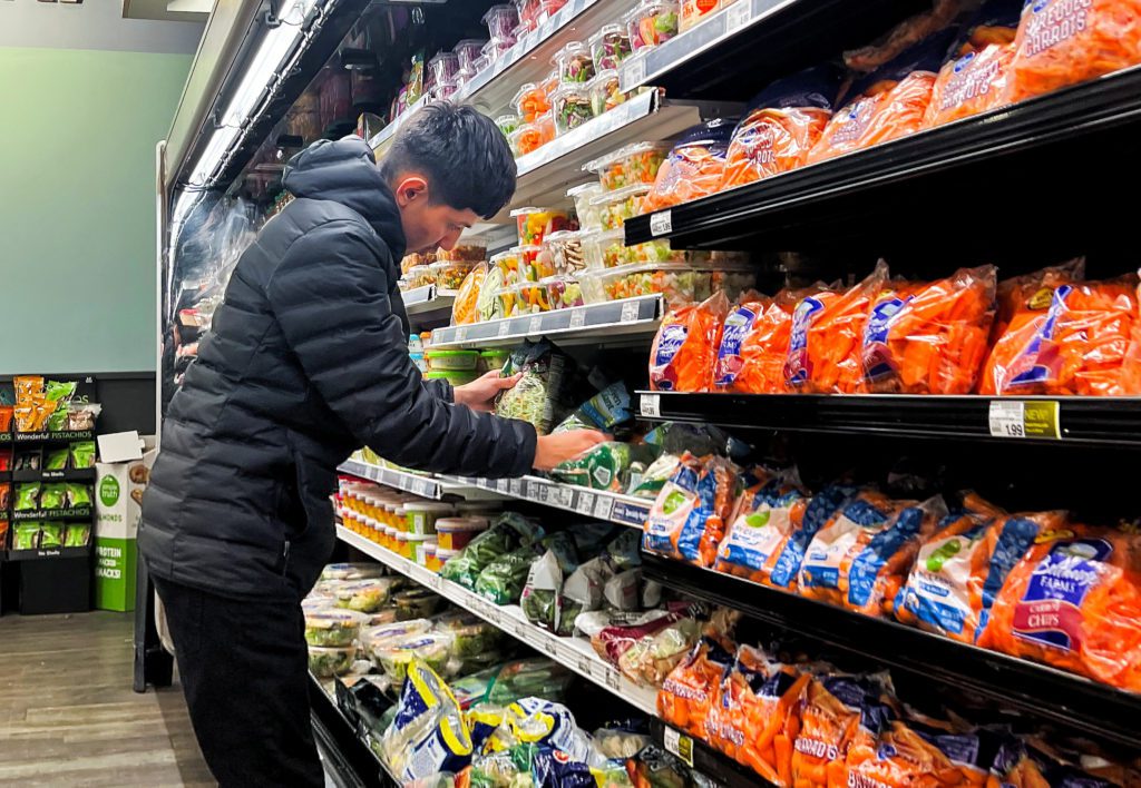 Homem escolhendo legumes embalados em prateleira refrigerada de mercado.