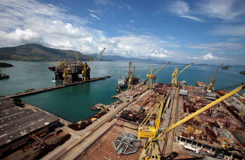 Estaleiro industrial com guindastes e plataforma offshore na água, e montanhas ao fundo sob céu azul.