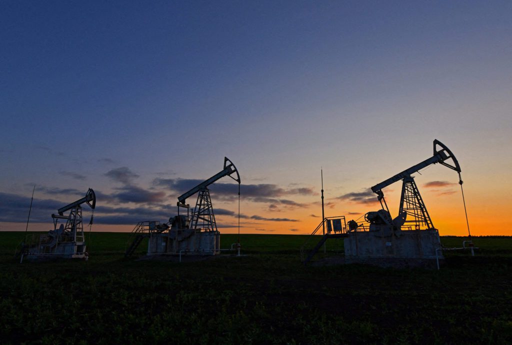 Três pumpjacks de petróleo em silhueta em campo. Entardecer com céu azul escuro e horizonte laranja.