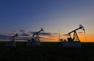 Três pumpjacks de petróleo em silhueta em campo. Entardecer com céu azul escuro e horizonte laranja.