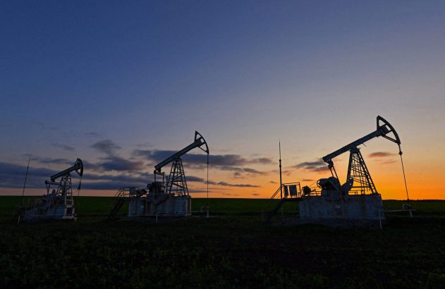 Três pumpjacks de petróleo em silhueta em campo. Entardecer com céu azul escuro e horizonte laranja.