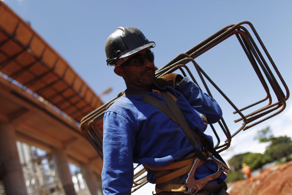 Trabalhador com capacete e uniforme azul carregando vergalhões em canteiro de obras.