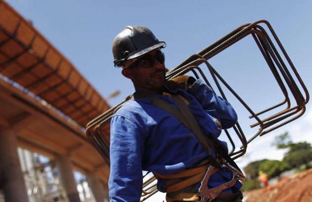 Trabalhador com capacete e uniforme azul carregando vergalhões em canteiro de obras.