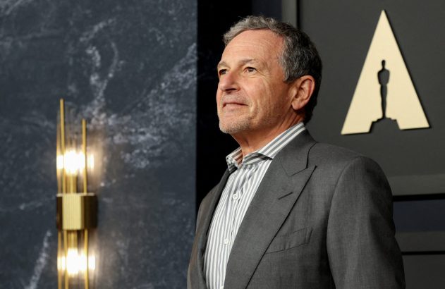 Homem de terno e camisa listrada, de perfil, olhando para a esquerda. Logo do Oscar e luminária ao fundo.