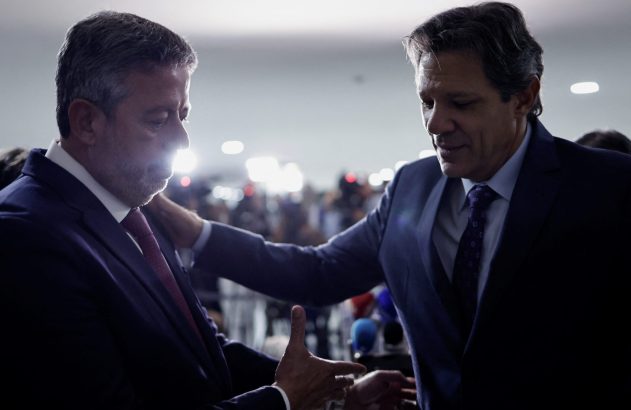 Dois homens de terno interagindo. Um coloca a mão no ombro do outro. Ambiente iluminado com contraluz e flashes.