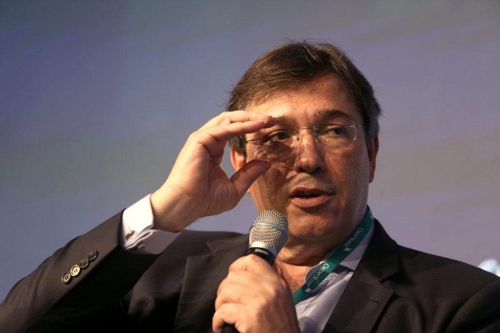 Homem de óculos, terno e crachá Credit Suisse, com microfone na mão, leva a outra mão ao rosto.