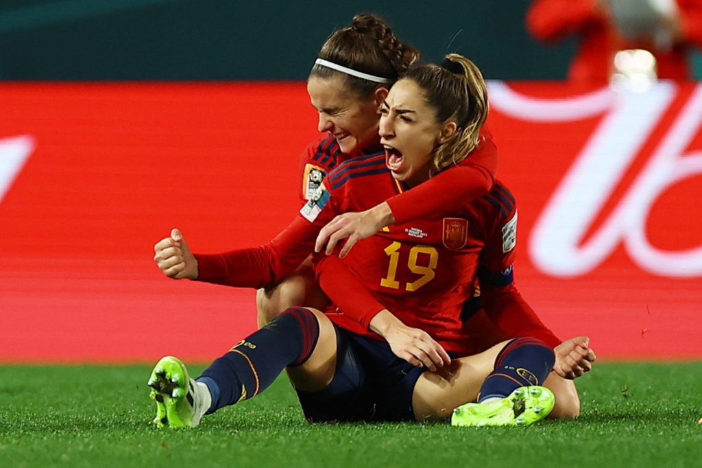 Duas jogadoras de futebol em uniformes vermelhos celebram intensamente no campo. Uma abraça a outra, que grita de alegria.