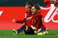 Duas jogadoras de futebol em uniformes vermelhos celebram intensamente no campo. Uma abraça a outra, que grita de alegria.
