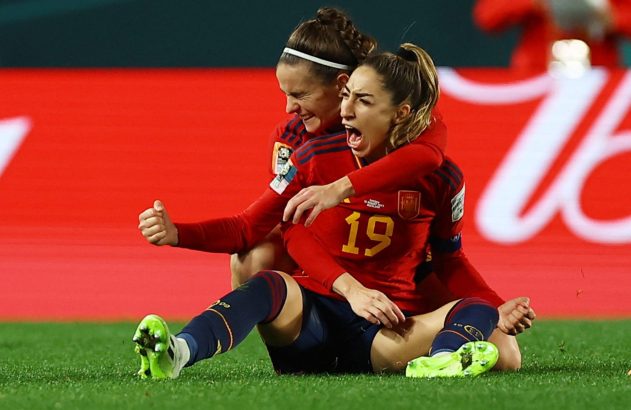 Duas jogadoras de futebol em uniformes vermelhos celebram intensamente no campo. Uma abraça a outra, que grita de alegria.