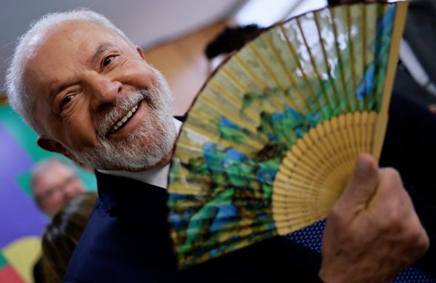 Homem de barba branca e cabelo grisalho sorri, segurando um leque colorido com desenhos de natureza.