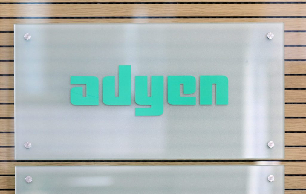 Placa de vidro fosco com o logo verde "adyen" fixada em parede de madeira clara.