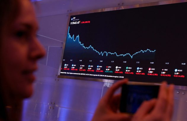 Mulher observa gráfico de queda em tela grande de dados financeiros, segurando um smartphone.