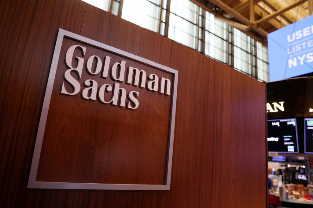 Logo Goldman Sachs em letras metálicas sobre painel de madeira. Fundo de escritório desfocado.