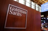 Logo Goldman Sachs em letras metálicas sobre painel de madeira. Fundo de escritório desfocado.