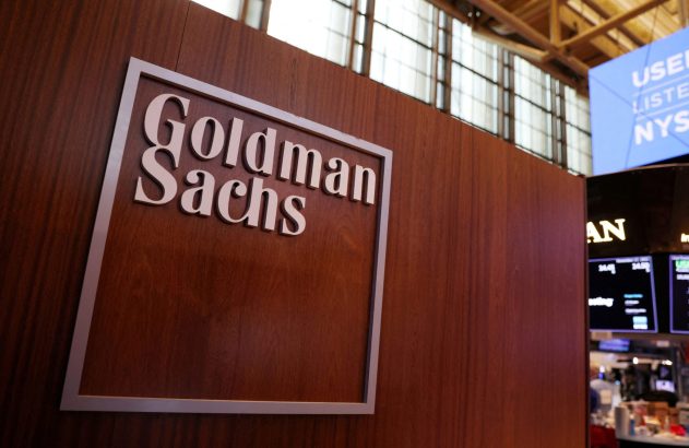 Logo Goldman Sachs em letras metálicas sobre painel de madeira. Fundo de escritório desfocado.