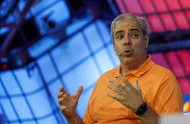 Palestrante de polo laranja gesticula ao falar, com fundo de luzes azuis e vermelhas.