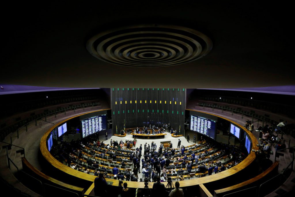 Grande plenário circular com parlamentares em sessão, monitores exibindo dados e equipe de mídia.