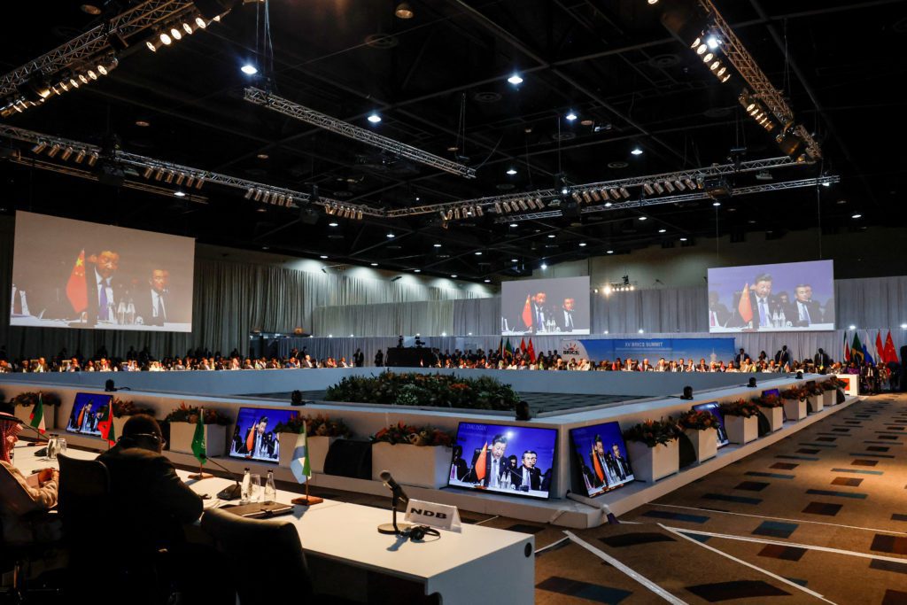 Participantes em uma cúpula do BRICS, com grandes telas projetando oradores e monitores individuais nas mesas.