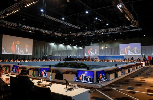 Participantes em uma cúpula do BRICS, com grandes telas projetando oradores e monitores individuais nas mesas.