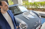 Homem de jaqueta azul e barba olha um Porsche cinza em ambiente interno.