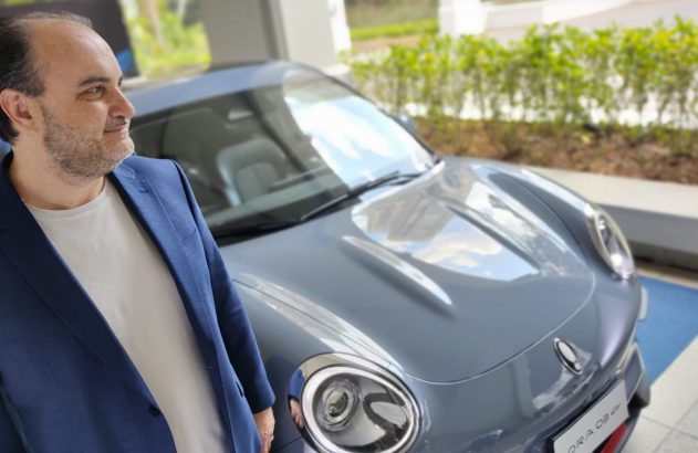 Homem de jaqueta azul e barba olha um Porsche cinza em ambiente interno.