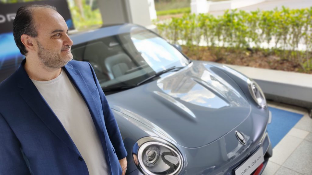 Homem em blazer azul ao lado de um carro cinza ORA 03 GT, olhando para o lado.