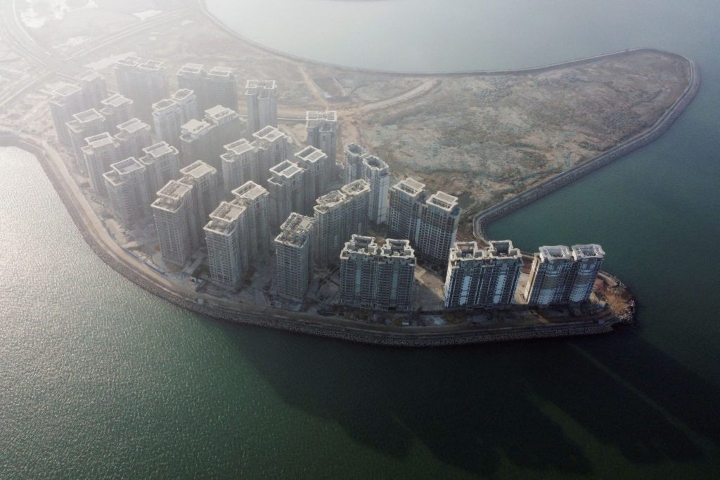 Vista aérea de uma ilha artificial com um grande complexo de edifícios altos, cercada por água esverdeada.