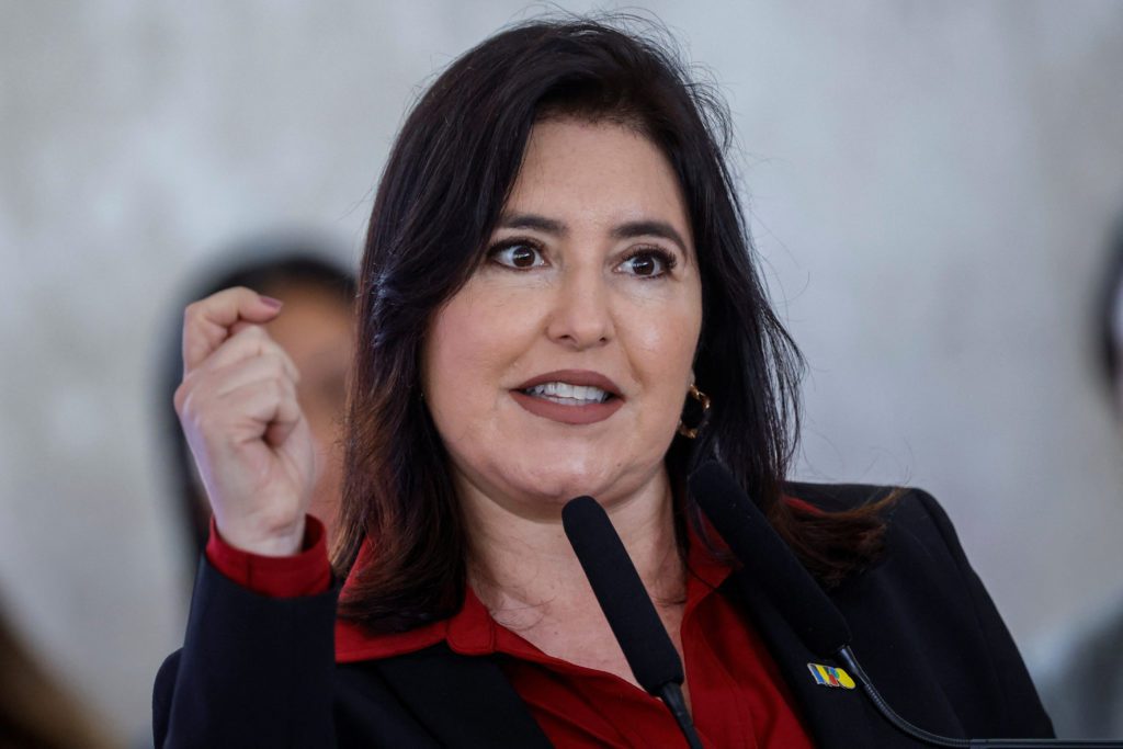 Mulher de cabelo escuro, camisa vermelha e blazer, fala em microfone e gesticula com o punho cerrado.