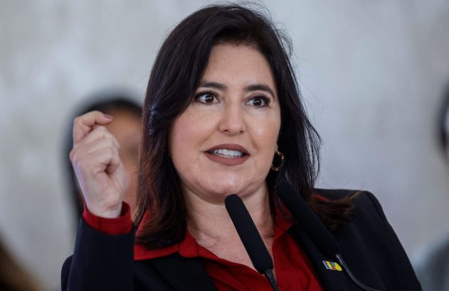 Mulher de cabelo escuro, camisa vermelha e blazer, fala em microfone e gesticula com o punho cerrado.