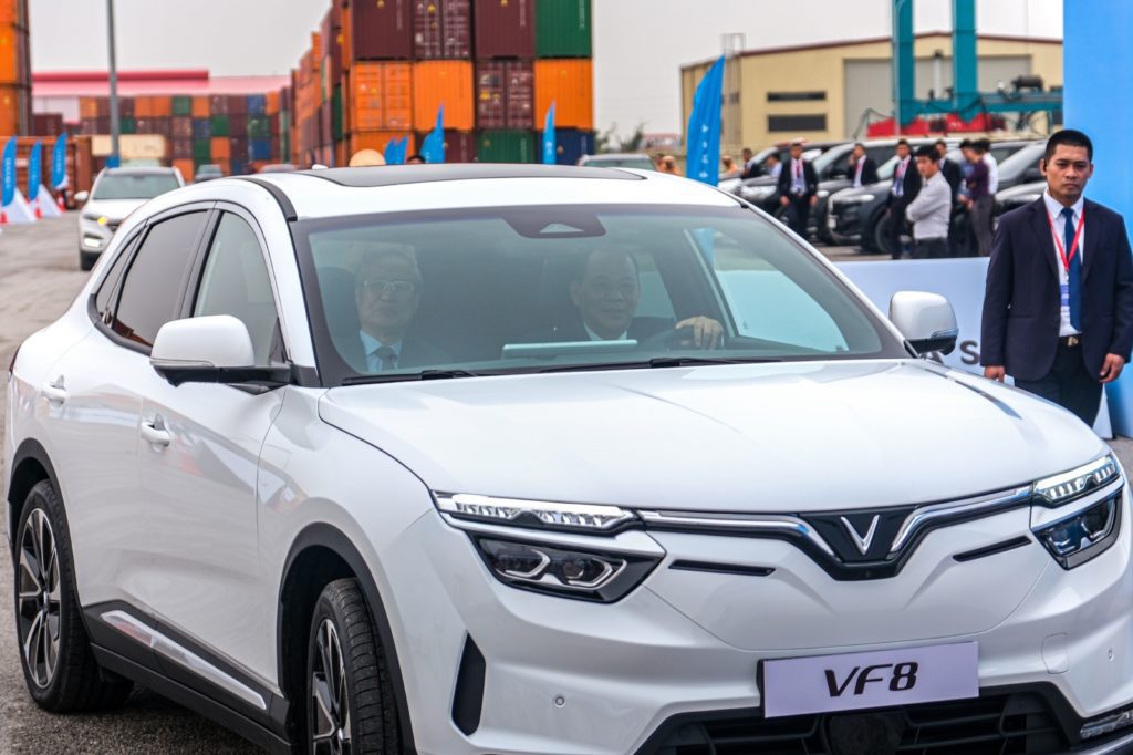 SUV VinFast VF8 branco sendo apresentado: pessoas dentro, homem ao lado, e contêineres de carga ao fundo.