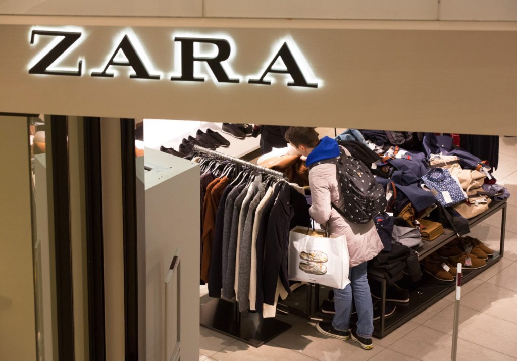 Vista aérea de cliente em loja Zara, olhando roupas em cabideiro e segurando sacola de compras. Placa "ZARA" visível.