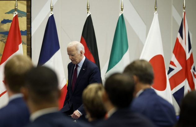 Joe Biden caminha à frente de bandeiras de vários países, com pessoas desfocadas em primeiro plano.