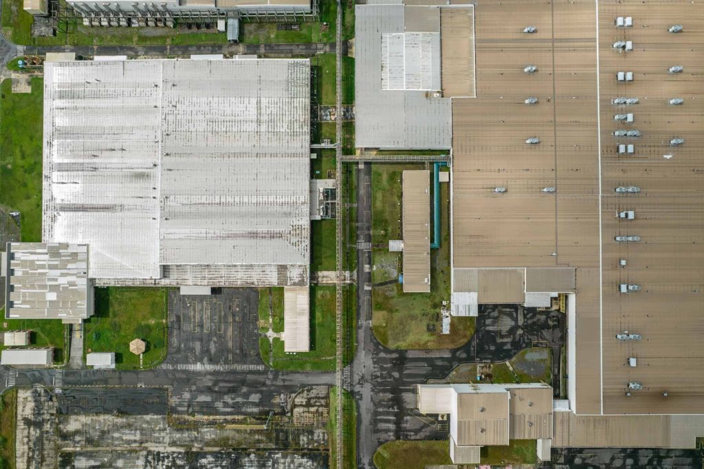 Vista aérea de um complexo industrial com grandes edifícios de telhados variados, estradas e áreas gramadas.