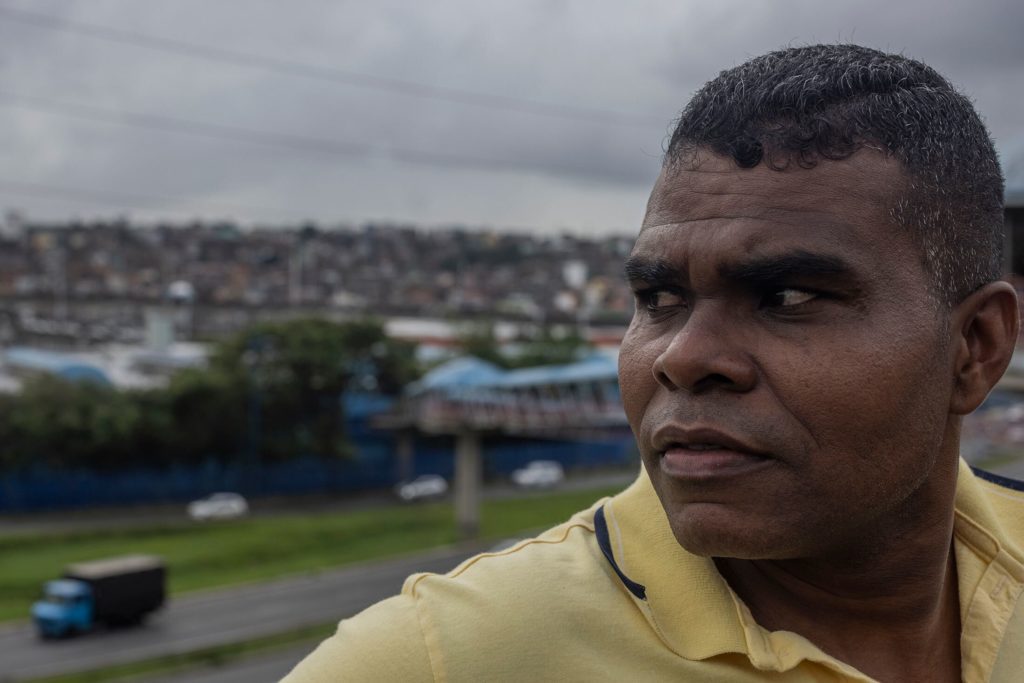 Homem de camisa amarela olha para o lado, com favela e rodovia ao fundo. Céu nublado.