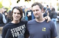 Dois homens sorrindo para a câmera. Um usa camisa "STUDENT FOR LIFE", o outro o abraça pelo ombro. Fundo com pessoas.