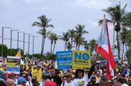 Manifestação com multidão, bandeira de Porto Rico e cartazes, incluindo um grande "NÃO".