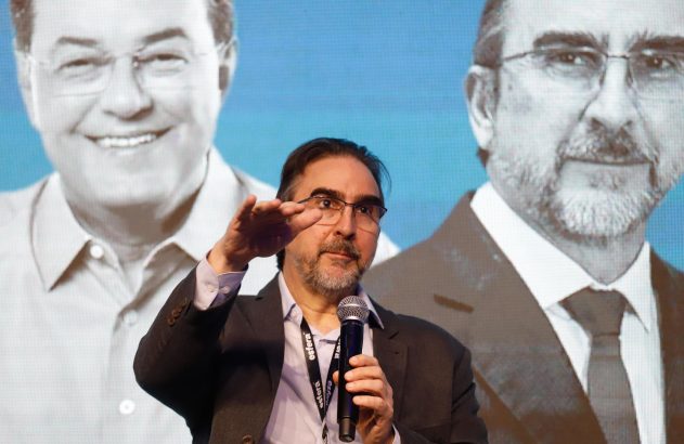 Palestrante com óculos e barba segura microfone e gesticula, à frente de imagens projetadas de dois homens.