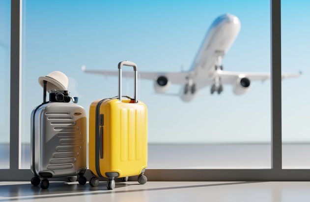 Duas malas (amarela e prata) em aeroporto. Avião decola pela janela, com céu azul ao fundo.