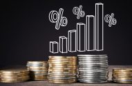Pilhas de moedas com gráfico de barras crescente e símbolos de porcentagem em fundo escuro, representando crescimento financeiro.