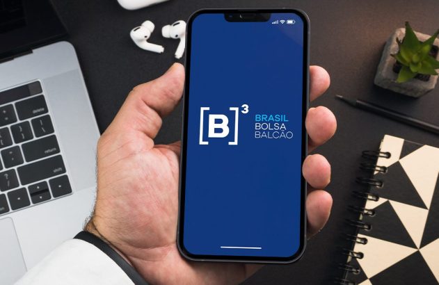 Mão segura celular com logo B3 Brasil Bolsa Balcão. Fundo: laptop, fones e caderno.