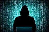 Hacker encapuzado em silhueta, usando laptop com fundo de código binário.