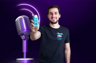 Homem sorridente apresenta bebida azul. Microfone e ondas sonoras roxas no fundo.