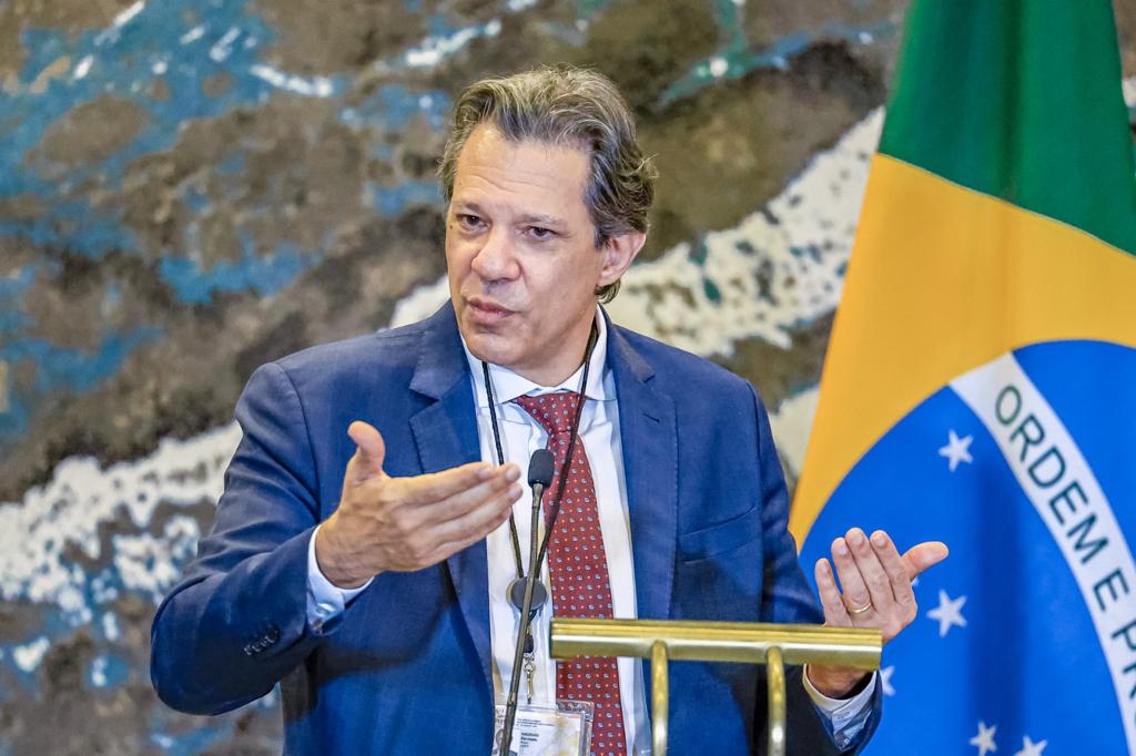 Homem de terno azul fala e gesticula. Bandeira do Brasil ao fundo.