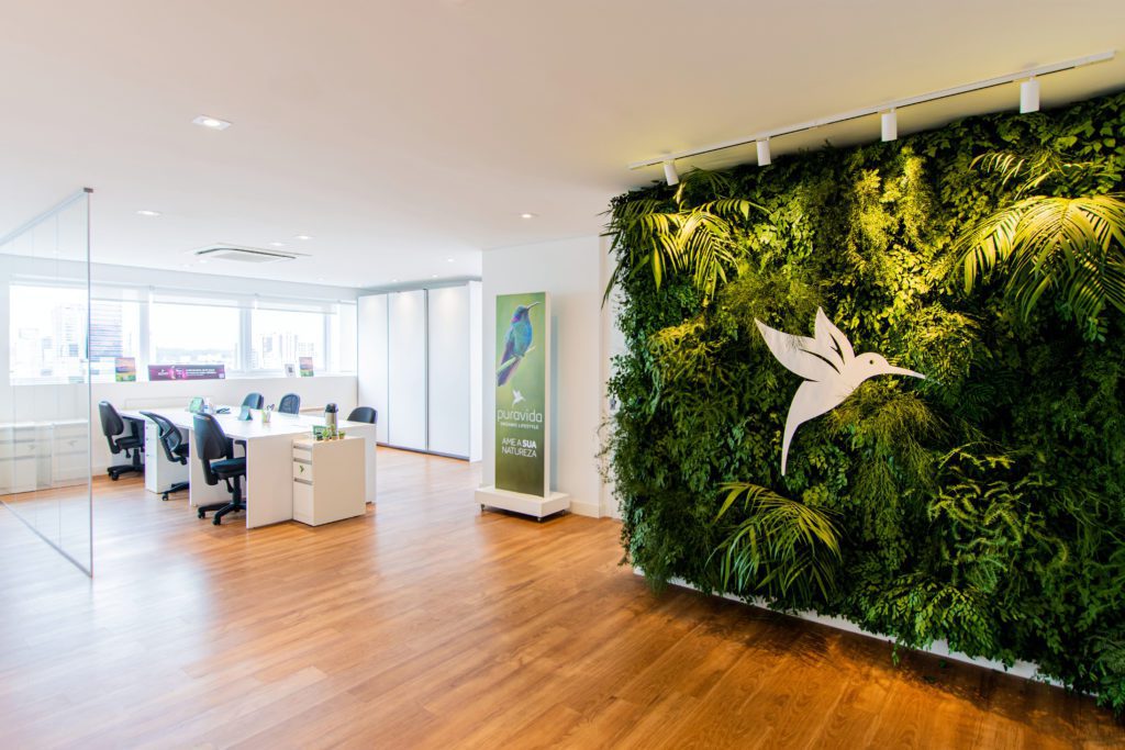 Escritório com jardim vertical grande, logo de colibri branco, e piso de madeira. Mesas e cadeiras ao fundo.