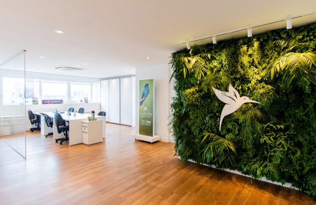 Escritório com jardim vertical grande, logo de colibri branco, e piso de madeira. Mesas e cadeiras ao fundo.