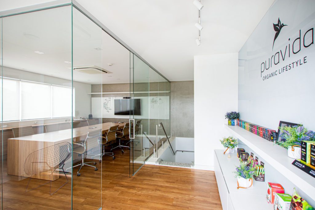 Interior de escritório moderno da PuraVida com sala de reunião de vidro, prateleiras de produtos e escada.