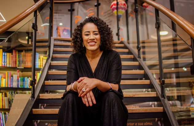 Mulher negra sorrindo sentada em escada de madeira, cercada por estantes de livros.